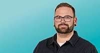 Mann mit Bart, Brille, Piercings vor türkisfarbenem Hintergrund