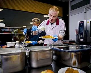 Zwei Personen mit Handschuhen geben Essen aus großen Behältern in einer Kantine