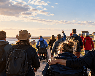 Gruppe von Menschen wandert auf einem Weg am Wasser mit einem Leuchtturm bei Sonnenuntergang