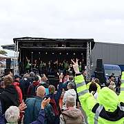 Menschenmenge vor einer kleinen Open-Air-Bühne mit Musikern und Veranstaltern bei bewölktem Himmel im Freien auf einem Industriehofgelände mit Fahrzeugen im Hintergrund, einige Zuschauer heben die Hände und zeigen Friedenszeichen oder jubeln; Personen auf