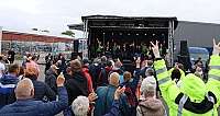 Menschenmenge vor einer kleinen Open-Air-Bühne mit Musikern und Veranstaltern bei bewölktem Himmel im Freien auf einem Industriehofgelände mit Fahrzeugen im Hintergrund, einige Zuschauer heben die Hände und zeigen Friedenszeichen oder jubeln; Personen auf