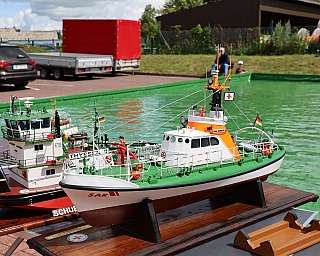 Mehrere detaillierte Modellboote in einem kleinen Wasserbecken im Freien mit parkenden Autos im Hintergrund und Menschen im Hintergrund auf Grünfläche sitzend. Das Vordergrundboot trägt eine deutsche Flagge und leuchtend orangefarbene Bereiche auf Deck un