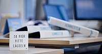 Holzschild mit Aufschrift auf Schreibtisch in Büro mit Monitoren im Hintergrund