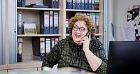 Frau mit Brille und lockigem Haar spricht am Telefon vor Aktenschrank