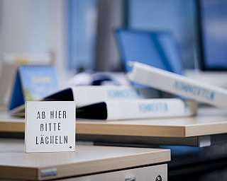 Schild auf Schreibtisch mit Aufschrift 'Ab hier bitte lächeln', Bürohintergrund