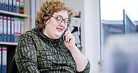 Frau mit Brille und lockigem Haar telefoniert lächelnd im Büro vor Aktenordnern