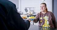 Frau mit braunem Pullover reicht einem anderen Menschen ein Teller mit Essen über einen Tresen