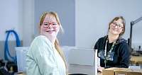 Zwei Frauen mit Brillen lächeln bei der Arbeit an einem Tisch mit Kartons