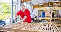 Mann in rotem Hemd bohrt Holz in Werkstatt mit weiteren Arbeitern und Holzstuhl