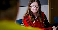 Frau mit Brille und rotem Sportjacke sitzt und spricht in einem Zimmer
