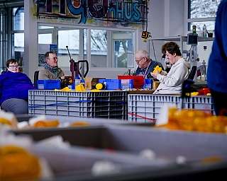 Mehrere Personen sitzen zusammen an Tischen mit gelben und blauen Werkstücken in Werkstatt