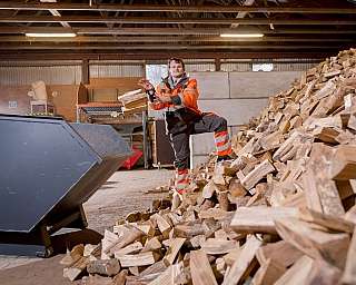 Mann in oranger Arbeitskleidung wirft Holzstücke in eine Maschine in einer Scheune mit großem Holzstapel daneben, Holzverarbeitungsszene in Halle mit Holzbalken und Steinboden, Innenaufnahme bei Tageslicht, Mülldeponie im Hintergrund sichtbar, Arbeitersic