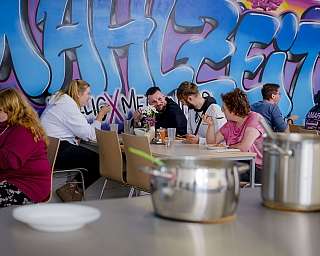 Gruppe von Menschen isst gemeinsam an Tischen vor bunter Graffiti-Wand