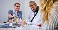 Drei Personen sitzen an einem Tisch mit Glasflasche und Dokumenten, lächeln