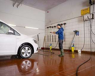 Mann in dunkler Kleidung wäscht weißen Volkswagen mit Hochdruckreiniger in Autowaschhalle